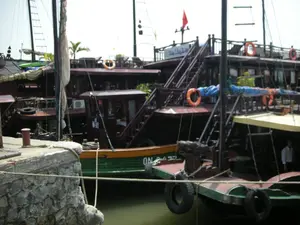 Bối cảnh là một bến tàu hoặc cầu cảng, có thể ở Vịnh Hạ Long, với nhiều tàu du lịch bằng gỗ đang neo đậu san sát nhau. Một cầu tàu bằng đá nằm ở phía trước bên trái, có dây thừng buộc các con tàu vào. Các con tàu này được thiết kế theo kiểu thuyền buồm truyền thống nhưng được trang bị các tiện nghi hiện đại; có thể thấy một cục nóng điều hòa không khí gắn bên ngoài một cabin. Trên boong tàu có các phao cứu sinh màu cam, và một chiếc thuyền kayak màu xanh lá cây được đặt trên nóc một con tàu. Mũi của một chiếc thuyền nhỏ ở phía trước được trang trí bằng một đầu rồng chạm khắc. Trên thân một con tàu có sơn màu xanh lá cây và cam, có ghi số hiệu "QN-3177". Trên tầng cao nhất của một con tàu khác có một tấm biển màu trắng nhưng chữ viết không thể đọc rõ, và một lá cờ đỏ đang bay. Một người đàn ông mặc áo sơ mi trắng đang đứng trên boong của một con tàu ở phía bên phải. Khung cảnh chung cho thấy các con tàu đang neo đậu tại bến, có thể đang chuẩn bị hoặc vừa kết thúc một chuyến tham quan.。（日本語訳）。背景は船着き場か埠頭で、おそらくハロン湾にあり、多くの木造の観光船が互いに密集して停泊している。左手前に石造りの桟橋があり、船を繋ぎ留めるためのロープがある。これらの船は伝統的な帆船の様式で設計されているが、現代的な設備が備え付けられている。手前にある小さな船の船首は、彫刻された龍の頭で装飾されている。ある船の船体には緑とオレンジの色が塗られており、登録番号「QN-3177」が記されている。別の船の最上階には白い看板があるが、文字ははっきりと読み取れず、赤い旗がはためいている。右側の船の甲板には、白いシャツを着た男性が一人立っている。全体的な光景は、船が船着き場に停泊していることを示しており、おそらくツアーの準備中か、あるいは終えたところだろう。