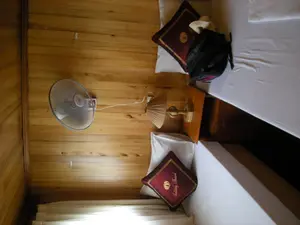 Bên trong một căn phòng có tường và sàn được ốp gỗ, có thể là phòng ngủ trên du thuyền. Trong phòng có hai giường đơn trải ga trắng. Trên mỗi giường có một chiếc gối tựa trang trí màu đỏ tía, một trong số đó có thêu dòng chữ "Halong Travel" và logo hình thuyền buồm. Giữa hai giường là một chiếc bàn gỗ nhỏ để đèn ngủ. Trên tường có gắn một chiếc quạt treo tường mang nhãn hiệu "VINASUN". Một chiếc ba lô màu đen được đặt trên một trong hai chiếc giường.。（日本語訳）。壁と床が木で張られた部屋の中。クルーズ船の寝室かもしれない。部屋には白いシーツが敷かれたシングルベッドが2つある。それぞれのベッドの上にはワインレッド色の装飾用クッションが1つあり、そのうちの1つには「Halong Travel」という文字と帆船のロゴが刺繍されている。2つのベッドの間には、ナイトランプを置くための小さな木製テーブルがある。壁には「VINASUN」ブランドの壁掛け扇風機が1台取り付けられている。