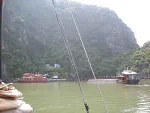 Bối cảnh là một vịnh, có thể là Vịnh Hạ Long, với những núi đá vôi đặc trưng được bao phủ bởi cây xanh rậm rạp. Dưới làn nước màu xanh lục, có một cầu tàu bằng bê tông nơi các tàu du lịch đang hoạt động. Một chiếc tàu du lịch lớn bằng gỗ, sơn màu nâu đỏ với mái ngói đỏ, đang neo đậu tại bến. Một chiếc tàu du lịch khác, cũng bằng gỗ màu nâu sẫm, đang ở gần cầu tàu, có thể đang trong quá trình cập bến hoặc rời đi. Trên cầu tàu có một nhóm người, dường như là du khách, đang đi bộ. Trên bờ, ở chân núi, có một vài công trình nhỏ, bao gồm một ngôi nhà mái xanh có treo cờ Việt Nam và một biển báo nhưng không thể đọc rõ nội dung. Khung cảnh được chụp từ một chiếc thuyền khác, với dây thừng của cột buồm ở phía trước.。（日本語訳）。背景は湾で、おそらくハロン湾であり、鬱蒼とした緑樹に覆われた特徴的な石灰岩の山々があります。緑色の水面には、観光船が運航しているコンクリート製の桟橋があります。赤い瓦屋根を持ち、赤褐色に塗装された大きな木造の観光船が、埠頭に停泊しています。もう一隻の観光船もまた濃い茶色の木造で、桟橋の近くにおり、接岸または離岸の最中である可能性があります。桟橋の上には、観光客と思われる一団の人々が歩いています。岸辺の、山の麓には、いくつかの小さな建造物があり、それにはベトナム国旗が掲げられた青い屋根の家と、内容ははっきりと読み取れない看板が含まれています。この光景は別の船から撮影されたもので、前方にはマストのロープがあります。