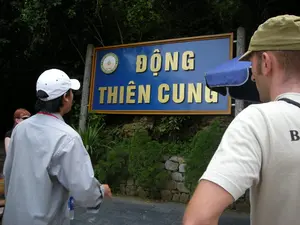 Bức ảnh chụp tại một khu vực ngoài trời, có vẻ là lối vào của một địa điểm du lịch hang động. Có một tấm biển hiệu lớn, nền xanh lam, viền gỗ, trên đó có dòng chữ nổi 3D màu vàng "ĐỘNG THIÊN CUNG". Ở góc trên bên trái của biển hiệu có một logo hình tròn ghi "DI SẢN THIÊN NHIÊN THẾ GIỚI" và "THE WORLD NATURAL HERITAGE".。Trong ảnh có ít nhất ba người, dường như là một nhóm du khách đang tham quan. Một người đàn ông mặc áo sơ mi xám, đội mũ lưỡi trai trắng đang nhìn về phía tấm biển. Một người đàn ông khác ở phía trước, mặc áo phông màu be và đội mũ màu xanh rêu, cũng đang nhìn vào biển hiệu. Phía sau họ, có thể thấy một phần của một người phụ nữ. Hoạt động của họ là đang xem biển chỉ dẫn trước khi vào hang động. Khung cảnh xung quanh có nhiều cây cối xanh tốt và một bức tường đá nhỏ, cho thấy đây là một khu vực tự nhiên được cải tạo để phục vụ du lịch.。（日本語訳）。写真は屋外で撮影されており、洞窟観光地の入口のようです。青い背景に木製の縁取りがされた大きな看板があり、その上には「ĐỘNG THIÊN CUNG」という黄色の3D立体文字が書かれています。看板の左上隅には、「DI SẢN THIÊN NHIÊN THẾ GIỚI」と「THE WORLD NATURAL HERITAGE」と書かれた円形のロゴがあります。写真には少なくとも3人の人物が写っており、観光中の旅行者グループのようです。灰色のシャツを着て白い野球帽をかぶった男性が、看板の方を見ています。その前にいる別の男性は、ベージュのTシャツを着てモスグリーンの帽子をかぶり、同じく看板を見ています。彼らの後ろには、女性の一部が見えます。彼らの行動は、洞窟に入る前に案内板を見ているところです。周囲の風景には青々とした木々や小さな石垣があり、ここが観光用に整備された自然エリアであることを示しています。