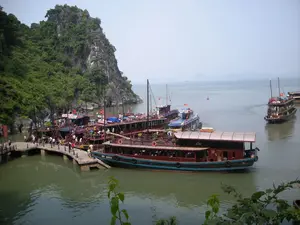 Bối cảnh là một bến tàu du lịch nhộn nhịp trên Vịnh Hạ Long, với nhiều tàu du lịch bằng gỗ đang neo đậu san sát tại một cầu tàu bê tông. Xung quanh là những hòn đảo đá vôi hùng vĩ đặc trưng của vịnh, được cây cối xanh tươi bao phủ. Rất đông người, chủ yếu là du khách, đang tập trung trên cầu tàu và trên các con tàu, thực hiện hoạt động lên và xuống tàu để tham quan vịnh. Một chiếc tàu du lịch bằng gỗ ở gần nhất có ghi số hiệu "QN 5985". Trên một chiếc tàu khác đang cập bến có biển hiệu ghi tên "BAI THO". Một số tàu có cắm cờ đỏ sao vàng của Việt Nam. Ở phía xa bên phải, một chiếc tàu khác đang di chuyển trên mặt biển.。（日本語訳）。背景はハロン湾の賑やかな観光船の船着き場で、多くの木造観光船がコンクリートの桟橋に密集して停泊しています。周りには、湾の特徴である雄大な石灰岩の島々が青々とした木々に覆われています。主に観光客である大勢の人々が桟橋や船の上に集まっており、湾を観光するために乗船・下船を行っています。最も近くにある木造観光船には「QN 5985」という船体番号が書かれています。接岸している別の船には、「BAI THO」という名前が書かれた看板があります。いくつかの船には、ベトナムの金星紅旗が掲げられています。右手の遠くでは、別の船が海上を移動しています。