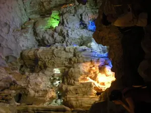 Bối cảnh là bên trong một hang động đá vôi, có thể là một điểm tham quan du lịch ở khu vực Vịnh Hạ Long. Các vách đá và các khối thạch nhũ, măng đá được chiếu sáng bởi hệ thống đèn màu nhân tạo, tạo ra các mảng màu sắc rực rỡ như xanh lá cây, xanh dương và vàng cam, làm nổi bật các hình thù kỳ lạ của đá. Ở phía bên phải của bức ảnh, có bóng của một vài du khách đang tham quan. Một người dường như đang giơ một vật giống máy ảnh lên để chụp lại cảnh quan trong hang.。（日本語訳）。背景は石灰岩の洞窟の中であり、ハロン湾地域の観光名所である可能性があります。岩壁や鍾乳石、石筍は人工的なカラー照明システムによって照らされ、緑、青、オレンジがかった黄色などの鮮やかな色彩を生み出し、奇妙な岩の形を際立たせています。写真の右側には、見学している数人の観光客の影があります。一人は、洞窟内の景色を撮影するためにカメラのようなものを掲げているようです。