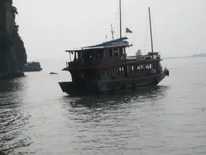 Bức ảnh chụp cảnh trên một vùng vịnh, có khả năng là Vịnh Hạ Long, với mặt nước phẳng lặng và bầu trời mờ sương. Nổi bật ở trung tâm là một chiếc tàu du lịch lớn, hai tầng, làm bằng gỗ. Thân tàu có màu sẫm với một sọc màu cam, và phần cabin trên có màu nâu. Trên nóc tàu có cột buồm và một lá cờ đỏ đang bay. Có thể thấy một vài người đang ở trên boong tàu. Con tàu này dường như đang di chuyển chậm trên mặt nước. Ở phía xa bên trái, gần một vách đá lớn, có một chiếc tàu khác tương tự. Khung cảnh cho thấy một hoạt động du lịch, tham quan vịnh bằng tàu. Không có chữ viết nào có thể đọc được trong ảnh.。（日本語訳）。写真は、おそらくハロン湾と思われる湾の風景を撮影したもので、水面は穏やかで、空は霧がかかっている。中央に目立っているのは、木造の大きな2階建ての観光船である。船体は暗い色でオレンジ色の縞模様があり、上部の船室は茶色である。船の屋根にはマストがあり、赤い旗がはためいている。この船は水面をゆっくりと移動しているようだ。左手の遠く、大きな崖の近くに、もう一隻の同様の船がある。この光景は、船で湾を観光する旅行活動を示している。写真の中には読み取れる文字はない。