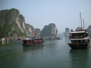 Đây là khung cảnh trên một vịnh biển, có thể là Vịnh Hạ Long, với nhiều hòn đảo đá vôi đặc trưng nhô lên từ mặt nước. Ở giữa và phía trước có hai chiếc tàu du lịch (tàu gỗ) lớn. Chiếc tàu bên trái có thân màu nâu đỏ và xanh, đang di chuyển và có cắm một lá cờ Việt Nam. Chiếc tàu bên phải có màu sẫm, trên tầng hai của tàu có một vài người dường như là du khách đang ngồi ngắm cảnh. Ở phía sau là một làng chài nổi với nhiều ngôi nhà và lồng bè được xây dựng trên mặt nước, cho thấy hoạt động sinh sống và có thể là nuôi trồng thủy sản của người dân địa phương. Hoạt động chính được ghi lại là du lịch tham quan vịnh bằng tàu thuyền.。（日本語訳）。これは湾の風景で、おそらくハロン湾でしょう。水面から突き出た多くの特徴的な石灰岩の島々があります。中央と手前には、2隻の大きな観光船（木造船）があります。左側の船は船体が赤褐色と緑色で、移動しており、ベトナムの国旗が一本掲げられています。右側の船は濃い色をしており、船の2階には観光客と思われる数人が座って景色を眺めています。後方には、水上に建てられた多くの家やいかだがある水上漁村があり、地元の人々の生活や、おそらくは水産養殖の営みを示しています。記録されている主な活動は、船による湾の観光ツアーです。