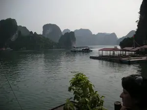 Bức ảnh ghi lại khung cảnh trên Vịnh Hạ Long, đặc trưng bởi những hòn đảo đá vôi trùng điệp nhô lên trên mặt nước lặng. Ở giữa và bên phải của bức ảnh là một khu nhà nổi lớn, có thể là một bè nuôi cá kết hợp dịch vụ du lịch hoặc một làng chài. Công trình này có nhiều gian với mái che màu đỏ, bên dưới có thể thấy một vài người và bàn ghế. Một lá cờ Việt Nam được cắm trên một cột cao. Ở phía xa hơn, có những chiếc thuyền và các nhà nổi khác nằm gần chân núi đá. Bầu trời có sương mù nhẹ, tạo nên một không gian yên tĩnh. Ở góc dưới bên phải, một người phụ nữ đang ngồi trên thuyền và ngắm nhìn cảnh vật. Không có văn bản nào có thể đọc được trong ảnh.。（日本語訳）。写真はハロン湾の風景を記録しており、穏やかな水面から突き出た無数の石灰岩の島々が特徴です。写真の中央と右側には大きな水上家屋群があり、それは観光サービスを兼ねた養殖いかだ、あるいは水上集落である可能性があります。この建造物には赤い屋根を持つ多くの区画があり、その下には数人の人々とテーブルや椅子が見えます。高いポールにベトナムの国旗が掲げられています。さらに遠くには、岩山の麓近くに船や他の水上家屋があります。空には薄い霧がかかっており、静かな空間を作り出しています。右下の角では、女性が船に座って景色を眺めています。写真の中には読み取れる文字はありません。