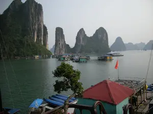 Bối cảnh là một làng chài nổi trên Vịnh Hạ Long, được bao quanh bởi những hòn đảo đá vôi hùng vĩ đặc trưng. Không có văn bản hay chữ viết nào có thể nhận dạng được. Hình ảnh cho thấy nhiều ngôi nhà nổi và bè nuôi cá với nhiều màu sắc khác nhau như xanh lam, xanh lá cây và vàng. Mái của một ngôi nhà ở phía trước có màu đỏ. Một lá cờ đỏ của Việt Nam được cắm trên một trong những nhà bè. Có nhiều chiếc thuyền kayak màu xanh được xếp gọn gàng, cho thấy sự hiện diện của các hoạt động du lịch. Cuộc sống hàng ngày của người dân làng chài diễn ra trên mặt nước, có khả năng bao gồm cả việc nuôi trồng thủy sản bên dưới các nhà bè. Một người đội mũ trắng có thể được nhìn thấy trên một trong những cấu trúc nổi.。（日本語訳）。背景はハロン湾に浮かぶ水上漁村で、特徴的な雄大な石灰岩の島々に囲まれています。認識できるテキストや文字はありません。画像には、青、緑、黄色など、さまざまな色の多くの水上家屋や魚の養殖いかだが見られます。手前にある家の屋根は赤色です。いかだの一つにベトナムの赤い国旗が掲げられています。青いカヤックが多数きれいに並べられており、観光活動の存在を示しています。漁村の人々の日常生活は水上で行われ、いかだの下での水産養殖も含まれている可能性があります。水上建造物の一つに、白い帽子をかぶった人が見えます。