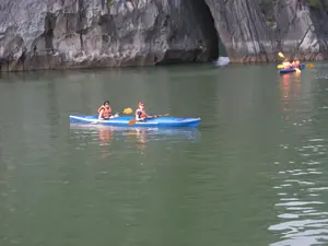 Bối cảnh là một khu vực trên mặt nước, có khả năng là ở Vịnh Hạ Long, được bao quanh bởi các vách núi đá vôi hùng vĩ. Có một cổng hang tự nhiên xuyên qua núi đá ở phía sau. Hoạt động chính đang diễn ra là chèo thuyền kayak. Ở phía trước, có một chiếc thuyền kayak đôi màu xanh dương với hai người phụ nữ đang ngồi trên đó. Cả hai đều mặc áo phao màu cam và cầm mái chèo. Ở phía xa hơn, một chiếc thuyền kayak khác cũng có hai người đang chèo về phía cổng hang. Mặt nước khá lặng và có màu xanh lục. Không có thông tin văn bản nào có thể đọc được trong hình ảnh.。（日本語訳）。背景は水上の一帯で、おそらくハロン湾で、雄大な石灰岩の崖に囲まれています。後方には、岩山を貫く自然の洞窟の入り口があります。行われている主なアクティビティはカヤックです。手前には、青い二人乗りのカヤックがあり、二人の女性が乗っています。二人ともオレンジ色のライフジャケットを着用し、パドルを持っています。さらに遠くには、別のカヤックにも二人が乗っており、洞窟の入り口に向かって漕いでいます。水面はかなり穏やかで、緑色をしています。画像内には読み取れるテキスト情報はありません。