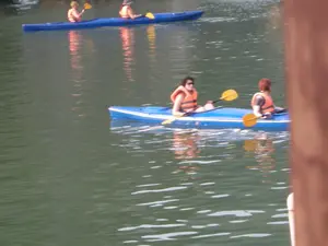 Bức ảnh chụp cảnh một nhóm người đang chèo thuyền kayak trên mặt nước yên tĩnh, có thể là ở một vịnh hoặc hồ lớn. Có hai chiếc thuyền kayak màu xanh lam. Chiếc thuyền ở gần hơn có hai người, cả hai đều mặc áo phao màu cam. Người ngồi phía trước có mái tóc sẫm màu và đeo kính râm. Người ngồi phía sau có mái tóc màu hung đỏ. Họ đang dùng mái chèo màu vàng để chèo thuyền. Ở phía xa hơn, có một chiếc thuyền kayak khác cũng có hai người. Họ cũng mặc áo phao màu cam và đội mũ. Đây là một hoạt động du lịch, giải trí chèo thuyền kayak. Mặt nước có màu xanh lục và phẳng lặng, phản chiếu hình ảnh của những người và thuyền. Không có chữ viết hay biển báo nào trong ảnh.。（日本語訳）。写真は、静かな水面でカヤックを漕いでいる人々のグループの光景を捉えています。おそらく湾か大きな湖でしょう。青いカヤックが2艘あります。手前にあるカヤックには2人が乗っており、2人ともオレンジ色のライフジャケットを着用しています。前に座っている人は黒っぽい髪で、サングラスをかけています。後ろに座っている人は赤みがかった髪色をしています。彼らは黄色いパドルを使ってボートを漕いでいます。さらに遠くには、もう1艘のカヤックがあり、こちらも2人が乗っています。彼らもオレンジ色のライフジャケットを着用し、帽子をかぶっています。これはカヤックを漕ぐ観光・レクリエーション活動です。水面は緑色で穏やかで、人々とボートの姿を映しています。写真には文字や標識はありません。