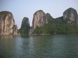 Bức ảnh chụp phong cảnh tại Vịnh Hạ Long. Nhiều hòn đảo đá vôi với các kích cỡ và hình dạng khác nhau nhô lên từ mặt biển. Các vách đá dựng đứng có màu xám và nâu, trong khi phần đỉnh và các sườn núi được bao phủ bởi thảm thực vật xanh tươi. Mặt nước biển có màu xanh đậm, gợn sóng nhẹ. Bầu trời có màu xanh nhạt và quang đãng. Không có sự hiện diện của con người hay tàu thuyền, tạo nên một khung cảnh thiên nhiên hùng vĩ và yên bình.。（日本語訳）。ハロン湾の風景を撮影した写真です。様々な大きさと形の石灰岩の島々が海面から突き出ています。切り立った崖は灰色と茶色で、頂上と山腹は青々とした植物に覆われています。海面は濃い青色で、穏やかに波立っています。空は薄い青色で、澄み渡っています。人や船の姿はなく、雄大で穏やかな自然の風景を作り出しています。