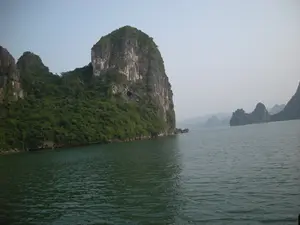 Đây là khung cảnh trên Vịnh Hạ Long. Bức ảnh chụp một hòn đảo đá vôi lớn sừng sững giữa mặt biển. Hòn đảo được bao phủ bởi thảm thực vật xanh tươi, đặc biệt là ở các sườn dốc, trong khi các vách đá dựng đứng lộ ra màu xám của đá. Có thể thấy một hang động nhỏ trên vách đá. Mặt nước biển có màu xanh ngọc bích đặc trưng, gợn sóng nhẹ. Góc chụp cho thấy bức ảnh được chụp từ một chiếc tàu du lịch đang di chuyển trên vịnh. Ở phía xa, nhiều hòn đảo đá vôi khác mờ ảo trong sương. Bầu trời có màu sáng và hơi mờ. Không có chữ viết hay biển báo nào trong ảnh. Hoạt động chính là du ngoạn, ngắm cảnh.。（日本語訳）。これはハロン湾の光景です。写真は、海の中央にそびえ立つ大きな石灰岩の島を写しています。島は、特に斜面が青々とした植物で覆われていますが、切り立った崖は岩の灰色を露出させています。崖には小さな洞窟が見えます。海面は特徴的なエメラルドグリーン色で、穏やかに波立っています。撮影角度から、写真が湾内を移動中の観光船から撮影されたことがわかります。遠くには、他の多くの石灰岩の島々が霧の中にぼんやりと見えます。空は明るく、少し霞んでいます。写真には文字や看板はありません。主なアクティビティは遊覧、景色を眺めることです。