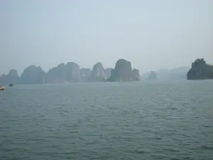 Bức ảnh chụp lại khung cảnh Vịnh Hạ Long. Toàn cảnh là một vùng biển rộng lớn, mặt nước gợn sóng nhẹ. Phía xa là vô số các hòn đảo đá vôi lớn nhỏ, nhấp nhô trên mặt biển, một đặc trưng của Vịnh Hạ Long. Bầu trời có sương mù bao phủ, khiến cho những hòn đảo ở xa trông mờ ảo. Ở phía bên trái, có một chiếc tàu du lịch màu đỏ đang di chuyển. Không có chữ viết hay văn bản nào có thể đọc được trong ảnh.。（日本語訳）。ハロン湾の風景を撮影した写真です。全景は広大な海で、水面は穏やかに波立っています。遠方には、ハロン湾の特徴である大小無数の石灰岩の島々が海面に突き出ています。空は霧に覆われており、遠くの島々が幻想的に見えます。左側には、赤い遊覧船が1隻、移動しています。写真の中には、読み取れる文字やテキストはありません。