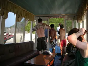 Bối cảnh là bên trong một chiếc tàu du lịch, có thể đang hoạt động ở khu vực Vịnh Hạ Long. Trên cửa ra vào có dòng chữ tiếng Anh "SEE YOU AGAIN". Một nhóm du khách, có vẻ là người nước ngoài, đang ở trên tàu. Một người đàn ông đang đứng và cởi chiếc áo phông sọc của mình. Gần đó, một người đàn ông khác cởi trần và một phụ nữ mặc đồ bơi đang đứng gần cửa ra vào, cho thấy họ có thể vừa trở về tàu sau khi bơi lội hoặc tham quan một bãi biển. Nội thất của tàu có ghế dài bọc da màu nâu, bàn gỗ và rèm cửa. Nhìn qua cửa sổ, có thể thấy một bãi biển cát trắng, những hòn đảo đá vôi với cây cối xanh tươi và một phần của một chiếc thuyền khác.。（日本語訳）。背景は、ハロン湾地域で運航している可能性のある観光船の船内です。出入り口には「SEE YOU AGAIN」という英語の文字があります。外国人と思われる観光客のグループが船に乗っています。一人の男性が立って、縞模様のTシャツを脱いでいます。その近くでは、上半身裸の別の男性と水着を着た女性が出入り口の近くに立っており、彼らが水泳やビーチ観光から船に戻ってきたばかりであることを示唆しています。船内には、茶色の革張りの長椅子、木製のテーブル、カーテンがあります。窓からは、白い砂浜、緑豊かな木々が生い茂る石灰岩の島々、そして別の船の一部が見えます。
