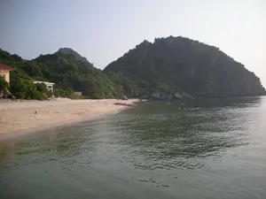 Bức ảnh chụp một khung cảnh bãi biển yên tĩnh, có thể là ở khu vực Vịnh Hạ Long. Cảnh vật bao gồm một bãi cát vàng ở bên trái, phía sau là những ngọn núi đá vôi được bao phủ bởi cây cối xanh tươi. Trên bãi biển có một vài ngôi nhà, trong đó có một ngôi nhà tường vàng, mái đỏ. Một vài chiếc thuyền nhỏ màu đỏ, có thể là thuyền kayak, đang đậu trên cát gần mép nước. Dưới biển, một nhóm người đang bơi lội. Mặt nước biển phẳng lặng và bầu trời có vẻ hơi mờ sương. Không có thông tin văn bản nào có thể nhận dạng được trong ảnh.。（日本語訳）。この写真は、おそらくハロン湾地域の、静かなビーチの風景を撮影したものです。風景には、左側に金色の砂浜があり、その後ろには青々とした木々に覆われた石灰岩の山々があります。ビーチにはいくつかの家があり、その中には黄色い壁と赤い屋根の家が1軒あります。おそらくカヤックと思われるいくつかの小さな赤いボートが、水際に近い砂の上に停泊しています。海では、数人のグループが泳いでいます。海面は穏やかで、空は少し霞んでいるようです。写真の中には、識別できるテキスト情報はありません。