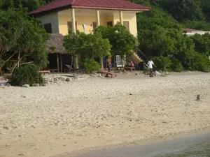 Bối cảnh là một bãi biển yên tĩnh, có thể nằm trên một hòn đảo ở khu vực Vịnh Hạ Long. Nổi bật trong ảnh là một ngôi nhà hai tầng sơn màu vàng với mái ngói đỏ, có hàng hiên với các cột trụ. Bên cạnh ngôi nhà chính là một công trình nhỏ hơn có mái lợp bằng lá, có thể là một quầy bar hoặc khu vực nghỉ ngơi. Trên bãi cát trắng có đặt một vài chiếc ghế dài để tắm nắng. Phía sau các công trình là cây cối xanh tươi, rậm rạp. Có hai người đàn ông trên bãi biển; một người mặc áo sơ mi trắng đang đi bộ trên cát, và người còn lại không mặc áo đang đứng gần khu vực nhà ở. Phía xa, có một tấm biển trên một tòa nhà khác nhưng chữ viết quá mờ để có thể đọc được.。（日本語訳）。背景は静かなビーチで、ハロン湾地域の島にあるのかもしれません。写真の中で目立っているのは、柱のあるベランダと赤い瓦屋根を持つ、黄色に塗られた2階建ての家です。母屋の隣には、葉で葺かれた屋根を持つ小さな建物があり、バーまたは休憩エリアかもしれません。白い砂浜の上には、日光浴用の長椅子がいくつか置かれています。建物の後ろには、青々とした木々が生い茂っています。ビーチには2人の男性がいます。白いシャツを着た一人は砂の上を歩いており、もう一人は上半身裸で住居エリアの近くに立っています。遠くには、別の建物に看板がありますが、文字がぼやけすぎていて読むことができません。