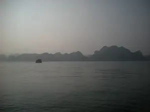 Bức ảnh chụp cảnh quan trên Vịnh Hạ Long. Khung cảnh bị bao phủ bởi một lớp sương mù dày đặc, tạo nên một không gian mờ ảo, tĩnh lặng. Ở phía xa, những dãy núi đá vôi đặc trưng của Vịnh Hạ Long hiện ra như những bóng đen trùng điệp. Trên mặt biển phẳng lặng, có một chiếc tàu (có thể là tàu du lịch) đang di chuyển. Toàn bộ cảnh vật mang một tông màu xám, gợi cảm giác yên bình và có phần huyền bí.。（日本語訳）。ハロン湾の風景を撮影した写真です。風景は厚い霧の層に覆われ、幻想的で静寂な空間を作り出しています。遠くには、ハロン湾特有の石灰岩の山々が、重なり合う黒い影のように現れています。穏やかな海の上を、一隻の船（おそらく観光船）が移動しています。全景は灰色のトーンを帯びており、平穏で、どこか神秘的な感覚を呼び起こします。