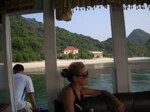 Bức ảnh được chụp từ bên trong một chiếc thuyền du lịch đang di chuyển trên mặt nước, có thể là ở khu vực vịnh. Qua khung cửa sổ của thuyền, có thể nhìn thấy một bãi biển cát trắng, phía sau là những ngọn núi đá vôi được cây cối bao phủ xanh tươi. Trên bờ biển có một vài công trình xây dựng, bao gồm một ngôi nhà màu vàng mái đỏ. Có một vài người đang ở trên bãi biển. Bên trong thuyền, một người phụ nữ đeo kính râm đang ngồi trên ghế và nhìn ra ngoài. Một người đàn ông mặc áo phông trắng đang ngồi ở lối đi mở của thuyền. Ở góc trên bên trái của bức ảnh, có một tấm biển dán trên cửa sổ với dòng chữ có thể đọc được là: '...ĐÃ ĐƯỢC BẢO HIỂM TẠI' và '...BẢO HIỂM DẦU KHÍ VIỆT NAM'.。（日本語訳）。写真は、水面を移動している観光船の内部から撮影されたもので、おそらく湾岸地域でしょう。船の窓枠からは、白い砂浜が見え、その後ろには緑豊かな木々に覆われた石灰岩の山々があります。海岸には、赤い屋根の黄色い家を含む、いくつかの建造物があります。ビーチには数人の人がいます。船内では、サングラスをかけた女性が椅子に座って外を眺めています。白いTシャツを着た男性が、船の開かれた通路に座っています。写真の左上隅には、窓に貼られたステッカーがあり、「...ĐÃ ĐƯỢC BẢO HIỂM TẠI」および「...BẢO HIỂM DẦU KHÍ VIỆT NAM」と読める文字があります。