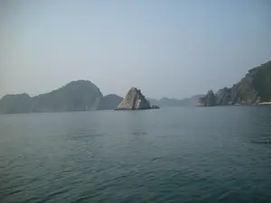 Đây là một khung cảnh trên Vịnh Hạ Long. Bức ảnh cho thấy một vùng biển rộng lớn với nhiều hòn đảo đá vôi có kích thước khác nhau nhô lên trên mặt nước. Các hòn đảo này được bao phủ bởi thảm thực vật xanh. Bầu trời có vẻ mờ ảo do sương mù, một đặc điểm thời tiết thường thấy ở khu vực này. Mặt biển tương đối phẳng lặng, chỉ có những gợn sóng nhỏ. Không có tàu thuyền hay hoạt động của con người nào có thể được nhìn thấy, chỉ có cảnh quan thiên nhiên hùng vĩ và tĩnh lặng.。（日本語訳）。これはハロン湾の風景です。これらの島々は緑の植生に覆われています。空は霧で霞んでいるように見えますが、これはこの地域でよく見られる天候の特徴です。海面は比較的に穏やかで、小さなさざ波しかありません。船や人間の活動は見られず、雄大で静かな自然の景観だけがあります。