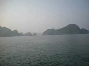 Bức ảnh chụp phong cảnh trên Vịnh Hạ Long. Khung cảnh chủ đạo là mặt biển rộng lớn, phẳng lặng với những gợn sóng nhỏ. Phía xa là vô số hòn đảo đá vôi lớn nhỏ, trùng điệp nhô lên trên mặt nước, đây là cảnh quan đặc trưng của khu vực này. Các hòn đảo được bao phủ bởi thảm thực vật xanh. Bầu trời có màu xám và bị bao phủ bởi một lớp sương mù dày đặc, khiến cho tầm nhìn xa bị hạn chế và tạo nên một không khí tĩnh lặng, mờ ảo. Ở khoảng cách xa, giữa các hòn đảo, có thể nhận ra một chiếc tàu đang di chuyển. Không có chữ viết hay ký hiệu nào trong ảnh.。（日本語訳）。ハロン湾で撮影された風景写真です。主な風景は、小さなさざ波が立つ、広大で穏やかな海面です。遠方には、大小無数の石灰岩の島々が重なり合って水面から突き出ており、これはこの地域の典型的な景観です。島々は緑の植生に覆われています。空は灰色で、厚い霧に覆われているため、遠くの視界が遮られ、静かで幻想的な雰囲気を醸し出しています。遠く、島々の間に、移動している一隻の船が認められます。写真には文字や記号はありません。