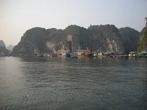Bức ảnh chụp một làng chài nổi trên mặt nước, có thể là ở Vịnh Hạ Long, vớiฉาก nền là những dãy núi đá vôi cao và hùng vĩ. Trên mặt nước có nhiều ngôi nhà nổi được xây dựng trên các bè phao, đây là nơi sinh sống và sinh hoạt của người dân. Nhiều loại tàu thuyền đang neo đậu san sát nhau, bao gồm các tàu đánh cá bằng gỗ cỡ lớn có kết cấu phức tạp và các thuyền nhỏ hơn. Một chiếc thuyền nhỏ màu xanh lam nổi bật ở giữa. Hoạt động chính ở đây dường như là đánh bắt và nuôi trồng thủy sản, thể hiện qua sự hiện diện của các tàu thuyền và cấu trúc của làng nổi. Không có văn bản hay chữ viết nào có thể nhận dạng được trong ảnh.。（日本語訳）。写真は水上に浮かぶ漁村を写しており、おそらくハロン湾にあるもので、背景には高く雄大な石灰岩の山々が連なっている。水上には、いかだの上に建てられた多くの水上家屋があり、ここは人々の生活の場となっている。複雑な構造を持つ大型の木造漁船やより小さなボートを含む、様々な種類の船が互いに密集して停泊している。中央には青い小さなボートが目立っている。ここでの主な活動は漁業と水産養殖であるようで、それは船の存在や水上村の構造から見て取れる。写真の中には識別できるテキストや文字はない。