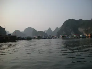 Bối cảnh là một làng chài nổi trên Vịnh Hạ Long, có thể là vào lúc sáng sớm hoặc chiều muộn do có lớp sương mù nhẹ. Ở trung tâm bức ảnh là một cụm các ngôi nhà nổi nhiều màu sắc, là nơi sinh sống của người dân. Xung quanh các ngôi nhà là các lồng bè, cho thấy hoạt động nuôi trồng thủy sản. Nhiều tàu thuyền, ghe nhỏ đang neo đậu gần các nhà nổi. Phía bên trái có một chiếc tàu gỗ lớn hơn, trên cột buồm có treo lá cờ Việt Nam. Phía sau làng chài là khung cảnh đặc trưng của Vịnh Hạ Long với những dãy núi đá vôi trùng điệp nhô lên từ mặt nước. Mặt biển khá lặng, chỉ có những gợn sóng nhỏ.。（日本語訳）。背景はハロン湾の水上漁村で、薄い霧がかかっていることから、早朝か夕方遅くかもしれません。写真の中央には、人々の生活の場である色とりどりの水上家屋の一群があります。家々の周りにはいけすがあり、水産養殖が行われていることを示しています。多くの船や小舟が水上家屋の近くに停泊しています。左側にはより大きな木造船があり、マストにはベトナム国旗が掲げられています。漁村の後ろには、水面から突き出る幾重にも連なる石灰岩の山々という、ハロン湾特有の風景が広がっています。海面はかなり穏やかで、小さな波紋があるだけです。