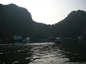 Quang cảnh một làng chài nổi trên mặt nước, có thể là ở Vịnh Hạ Long, vào thời điểm hoàng hôn hoặc bình minh. Bầu trời sáng nhưng không có mặt trời. Xung quanh là những hòn đảo và núi đá vôi cao, được bao phủ bởi cây xanh rậm rạp. Trên mặt nước, có nhiều ngôi nhà nổi và lồng bè. Một chiếc thuyền máy nhỏ (ghe) đang chạy trên mặt nước, để lại một vệt sóng phía sau. Có người đang ở trên thuyền. Ở phía xa hơn, có những chiếc tàu lớn hơn và các công trình nhà nổi khác, tạo nên một khung cảnh sinh hoạt của người dân sống trên biển.。（日本語訳）。水上漁村の光景、おそらくハロン湾で、夕暮れ時か夜明け時のものです。空は明るいですが、太陽は見えません。周りには、鬱蒼とした緑樹に覆われた高い島々や石灰岩の山々があります。水上には、多くの水上家屋やいけすがあります。一隻の小型モーターボート（ゲ）が水上を走り、後ろに航跡を残しています。船には人が乗っています。さらに遠くには、より大きな船や他の水上家屋があり、海上で暮らす人々の生活風景を創り出しています。