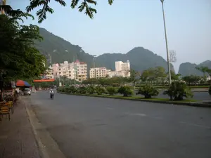 Trên vỉa hè bên trái, có một biển hiệu ghi chữ "CƠM", cho thấy đây là một quán ăn. Ở phía xa hơn, có một băng rôn màu đỏ treo ngang đường nhưng không thể đọc rõ chữ.。Bức ảnh chụp một con đường nhựa rộng lớn tại một thành phố, có những ngọn núi đá vôi làm nền, đặc trưng cho cảnh quan khu vực Hạ Long. Con đường có dải phân cách ở giữa được trồng cây xanh như cây cọ. Bên lề đường bên trái là một quán ăn hoặc quán nước vỉa hè với bàn ghế sắt và một chiếc ô lớn. Một người đang điều khiển xe máy trên đường. Phía sau hàng cây là các tòa nhà nhiều tầng, có thể là khách sạn hoặc khu dân cư.。（日本語訳）。左側の歩道上には、「CƠM」と書かれた看板があり、ここが食堂であることを示しています。さらに遠くには、道を横切って赤い横断幕が掲げられていますが、文字ははっきりと読めません。この写真は、ハロン地域の景観に特徴的な石灰岩の山々を背景にした、ある都市の広大なアスファルトの道を写しています。道の中央には、ヤシの木のような緑の木が植えられた中央分離帯があります。左側の路肩には、鉄製のテーブルと椅子、そして大きなパラソルを備えた歩道の食堂またはカフェがあります。一人の人が道路でバイクを運転しています。並木の背後には、ホテルまたは住宅地である可能性のある高層ビルがあります。