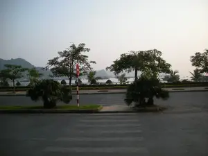 Bức ảnh chụp một con đường ven biển vào một thời điểm yên tĩnh, có thể là sáng sớm hoặc chiều muộn. Ở tiền cảnh là một con đường nhựa có vạch kẻ dành cho người đi bộ. Giữa đường có một dải phân cách nhỏ trồng cỏ và các bụi cây cọ lùn. Một cột biển báo giao thông sơn sọc đỏ trắng được cắm ở dải phân cách, trên đó có biển báo hình tròn nền đỏ với một vạch trắng ngang ở giữa, đây là biển báo 'Cấm đi ngược chiều'. Phía bên kia đường là vỉa hè trồng nhiều cây xanh như cây phượng và cây cọ. Xa hơn là khung cảnh của một vịnh biển, có thể là Vịnh Hạ Long, với mặt nước phẳng lặng. Trên vịnh có rất nhiều tàu thuyền, bao gồm cả tàu du lịch lớn và các thuyền nhỏ hơn, đang neo đậu. Ở phía xa, những dãy núi đá vôi đặc trưng nhô lên trên mặt nước, chìm trong một lớp sương mờ nhạt. Bầu trời có màu sáng nhưng không trong xanh, tạo cảm giác thanh bình và tĩnh lặng.。（日本語訳）。写真は、静かな時間帯、おそらく早朝か夕方遅くに撮影された海沿いの道を写している。前景には、歩行者用の横断歩道があるアスファルトの道がある。道の中央には、草と低いヤシの木の茂みが植えられた小さな中央分離帯がある。中央分離帯には赤と白の縞模様に塗られた交通標識のポールが立てられており、その上には中央に白い横線が一本入った赤地の円形の標識がある。これは「進入禁止」の標識である。道の向こう側は、ホウオウボクやヤシの木のような多くの緑の木が植えられた歩道である。さらに遠くには、穏やかな水面を持つ海の湾の風景が広がっており、ハロン湾かもしれない。湾には、大きな観光船やより小さなボートを含む多くの船が停泊している。遠方では、特徴的な石灰岩の山々が水面から突き出ており、薄い霧の中に沈んでいる。空は明るいが青く澄んではおらず、平和で静寂な感覚を生み出している。