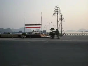 Trong ảnh có một biểu ngữ lớn với dòng chữ "CÁT BÀ KÍNH CHÀO QUÝ KHÁCH". Bên dưới là một băng rôn màu đỏ với chữ vàng, có thể là một khẩu hiệu chính trị.。Khung cảnh là một khu vực công cộng ven biển, có thể là quảng trường hoặc đường đi dạo ở Cát Bà, thuộc khu vực Vịnh Hạ Long. Phía trước là một con đường nhựa rộng. Phía sau con đường là một không gian mở được lát gạch với các cột đèn trang trí bằng kim loại có hình dạng giống cây dừa, một vài chậu cây cảnh bonsai và một hàng rào lan can nhìn ra biển.。Ở phía sau, có thể thấy một bến cảng với rất nhiều tàu thuyền đang neo đậu. Xa hơn nữa là mặt biển và những hòn đảo núi đá vôi đặc trưng của Vịnh Hạ Long. Khung cảnh khá yên tĩnh, có thể được chụp vào sáng sớm hoặc chiều muộn.。（日本語訳）。写真には「カットバは皆様を歓迎します」という文字が書かれた大きな横断幕があります。その下には黄色い文字の赤い横断幕があり、おそらく政治的なスローガンでしょう。風景は、ハロン湾地域に属するカットバ島の海辺の公共エリアで、広場か遊歩道かもしれません。手前には広いアスファルトの道路があります。道路の向こう側は、ヤシの木のような形をした金属製の装飾的な街灯柱、いくつかの盆栽の鉢植え、そして海を見渡す手すりの柵がある、タイル張りのオープンスペースです。後方には、多くの船が停泊している港が見えます。さらに遠くには、海面とハロン湾特有の石灰岩の島々があります。風景はかなり静かで、早朝か夕方遅くに撮影されたのかもしれません。