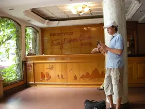 Bối cảnh là sảnh chờ và quầy lễ tân của một khách sạn. Trên bức tường gỗ lớn phía sau quầy có khắc dòng chữ 'RECEPTION', ba ngôi sao (***), và tên khách sạn 'Sunflower One HOTEL'. Một người đàn ông châu Á, đội mũ lưỡi trai trắng, mặc áo phông xanh nhạt và quần short, đang đứng dựa vào một chiếc cột lớn ốp đá cẩm thạch. Anh ta đang chăm chú sử dụng điện thoại di động. Dưới chân anh là một chiếc túi du lịch màu đen. Phía sau quầy lễ tân bằng gỗ, có thể thấy một nhân viên đang ngồi. Mặt trước của quầy được trang trí bằng các họa tiết chạm khắc bằng gỗ, mô tả những chiếc thuyền buồm và các hòn đảo núi đá, có thể là hình ảnh gợi nhớ đến Vịnh Hạ Long. Bên trái có các cửa sổ lớn nhìn ra cây cối xanh tốt bên ngoài.。（日本語訳）。背景はホテルの待合ロビーとフロントカウンターです。カウンターの後ろにある大きな木の壁には、「RECEPTION」の文字、3つの星（***）、そしてホテル名「Sunflower One HOTEL」が刻まれています。白い野球帽をかぶり、水色のTシャツとショートパンツを着用したアジア人男性が、大理石で覆われた大きな柱に寄りかかって立っています。彼は熱心に携帯電話を使っています。彼の足元には黒い旅行カバンがあります。木製のフロントカウンターの後ろには、座っているスタッフの姿が見えます。カウンターの前面は、帆船や岩山のような島々を描写した木彫りのモチーフで装飾されており、ハロン湾を彷彿とさせるイメージかもしれません。