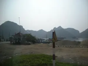 Bối cảnh là một bến cảng hoặc bến tàu ở một vùng ven biển, có khả năng là tại Vịnh Hạ Long, với những dãy núi đá vôi hùng vĩ đặc trưng làm nền trong làn sương mờ. Ở phía trước, có một biển báo giao thông hình tam giác trên cột sơn sọc đỏ trắng, cảnh báo một khúc cua nguy hiểm, nhưng không có chữ viết. Các chữ viết khác trên các tòa nhà hoặc tàu thuyền (nếu có) đều không thể đọc được do khoảng cách và chất lượng hình ảnh. Cảnh vật cho thấy một khu vực đất trống với các đống vật liệu trông giống như cát. Một trạm gác nhỏ có mái ngói nằm gần bến tàu, nơi một nhóm người và một chiếc xe buýt màu trắng đang tụ tập, có lẽ là khách du lịch đang chờ để lên thuyền. Nhiều tàu thuyền, bao gồm cả các tàu du lịch, đang neo đậu tại bến và một số khác đang di chuyển trên mặt vịnh. Một vài lá cờ đỏ sao vàng của Việt Nam có thể được nhìn thấy trên các con tàu. Hoạt động chính dường như là vận tải và du lịch đường thủy.。（日本語訳）。前景には、赤と白の縞模様に塗られた柱に三角形の交通標識があり、危険なカーブを警告しているが、文字は書かれていない。埠頭の近くに瓦屋根の小さな警備所があり、そこでは一群の人々と白いバスが集まっており、おそらく船に乗るのを待っている観光客だろう。観光船を含む多くの船が埠頭に停泊しており、その他は湾上を移動している。船の上には、ベトナムの金星紅旗がいくつか見られる。主な活動は水上輸送と観光のようである。