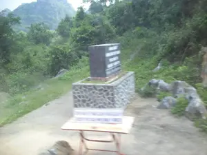 Trong ảnh là một bia tưởng niệm bằng đá đặt ven đường.。**Thông tin văn bản:** Có một tấm bia đá màu đen, trên đó có khắc chữ màu trắng nhưng không thể đọc được do ảnh mờ. Phía trước, có một chiếc bàn gấp nhỏ, trên bàn có một tấm biển khác nhưng chữ viết trên đó cũng bị mờ nhòe hoàn toàn do chuyển động.。**Mô tả cảnh vật và hoạt động:** Bối cảnh là một khu vực ven đường ở vùng nông thôn, miền núi với những ngọn núi đá vôi và cây cối rậm rạp ở phía sau. Vật thể chính là một bia đá tưởng niệm được đặt trên một bệ xây bằng đá. Bên cạnh là một con đường đất. Không có người xuất hiện và không có hoạt động nào đang diễn ra trong ảnh.。（日本語訳）。**テキスト情報：**黒い石碑があり、その上に白い文字が刻まれていますが、写真がぼやけているため読むことはできません。手前には小さな折りたたみテーブルがあり、テーブルの上には別の看板がありますが、その上の文字も動きによって完全にぼやけています。**風景と活動の説明：**背景は、後ろに石灰岩の山々と鬱蒼とした木々がある、田舎の山間部の道端の地域です。主な物体は、石で造られた台座の上に置かれた石の記念碑です。隣には未舗装の道があります。人は写っておらず、写真の中で行われている活動もありません。