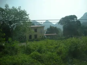 Bức ảnh chụp một khung cảnh ở vùng nông thôn. Nổi bật trong ảnh là một ngôi nhà hai tầng sơn màu vàng với mái ngói đỏ, trên mái có lắp một chiếc chảo vệ tinh. Bên cạnh ngôi nhà chính là một công trình phụ nhỏ hơn cũng có mái đỏ. Toàn bộ khu vực được bao quanh bởi cây cối xanh tươi và các bụi rậm mọc um tùm ở tiền cảnh. Ở phía sau, có thể thấy những dãy núi đá vôi mờ ảo trong sương. Một vài đường dây điện chạy ngang qua phần trên của bức ảnh. Không có người hay hoạt động cụ thể nào diễn ra. Không có chữ viết hay biển hiệu nào trong ảnh.。（日本語訳）。写真は田舎の風景を写しています。写真の中で目立っているのは、赤い瓦屋根の黄色い二階建ての家で、屋根には衛星放送受信アンテナが設置されています。母屋の隣には、同じく赤い屋根の小さな付属の建物があります。周辺一帯は緑豊かな木々に囲まれ、前景には茂った低木が生い茂っています。写真の上部を数本の電線が横切っています。人や特定の活動は見られません。写真には文字や看板はありません。