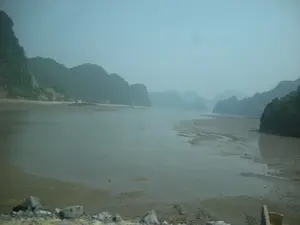 Bức ảnh ghi lại một khung cảnh ven biển, rất có thể là tại khu vực Vịnh Hạ Long, vào thời điểm thủy triều đang xuống thấp. Nước biển đã rút đi, để lộ ra một bãi sình lầy và cát rộng lớn ở giữa vịnh. Xung quanh là những ngọn núi đá vôi hùng vĩ, được bao phủ bởi cây cối xanh tươi, đặc trưng của vùng này. Bầu trời có sương mù dày đặc, khiến cho cảnh vật ở xa trở nên mờ ảo. Ở phía bên trái, có một con đường chạy dọc theo bờ biển dưới chân núi. Không có tàu thuyền hay hoạt động nào của con người được quan sát rõ, chỉ có một khung cảnh thiên nhiên tĩnh lặng. Phần tiền cảnh có những tảng đá, cho thấy góc chụp có thể là từ trên một con đường hoặc một điểm cao ven bờ.。（日本語訳）。写真は、おそらくハロン湾地域の、潮が引いている時の海岸の風景を記録しています。海水が引き、湾の中央に広大な泥と砂の干潟が現れています。周りには、この地域の特徴である、青々とした木々に覆われた雄大な石灰岩の山々があります。空には濃い霧がかかっており、遠くの景色がぼんやりと見えます。左側には、山の麓の海岸線に沿って走る道があります。船や人の活動ははっきりと観察されず、静かな自然の風景だけがあります。前景には岩があり、撮影アングルは道の上か、あるいは海岸沿いの高台からである可能性を示しています。