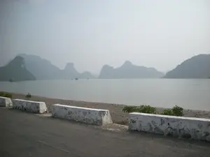 Bức ảnh chụp phong cảnh Vịnh Hạ Long nhìn từ một con đường ven biển. Ở phía trước là một con đường trải nhựa với các khối rào chắn bằng bê tông sơn trắng đã cũ. Phía sau rào chắn là một bãi sỏi nhỏ ven bờ. Trên mặt vịnh phẳng lặng, có một vài chiếc tàu du lịch đang di chuyển. Ở phía xa, nhiều hòn đảo đá vôi đặc trưng của Vịnh Hạ Long hiện ra mờ ảo trong sương mù dày đặc. Bầu trời xám và nhiều sương, tạo nên một không gian yên tĩnh.。（日本語訳）。海岸沿いの道から見たハロン湾の風景写真。手前は、古びた白いペンキが塗られたコンクリート製の防護ブロックがある舗装道路です。防護柵の後ろは、岸辺の小さな砂利浜です。穏やかな湾の水面には、数隻の遊覧船が移動しています。遠くには、ハロン湾特有の多くの石灰岩の島々が、濃い霧の中にぼんやりと現れています。空は灰色で霧が深く、静かな空間を作り出しています。