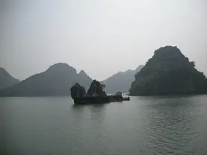 Đây là khung cảnh thiên nhiên tại Vịnh Hạ Long. Bức ảnh cho thấy một vùng biển rộng lớn với nhiều hòn đảo đá vôi có kích thước khác nhau nhô lên trên mặt nước. Bầu trời có sương mù nhẹ, khiến cho các hòn đảo ở phía xa trở nên mờ ảo. Các hòn đảo được bao phủ bởi thảm thực vật xanh. Ở trung tâm bức ảnh là một cụm đá nhỏ với hình dáng đặc biệt. Mặt nước phẳng lặng. Không có sự xuất hiện của con người, tàu thuyền hay bất kỳ hoạt động nào, chỉ có vẻ đẹp yên tĩnh và hùng vĩ của tự nhiên.。（日本語訳）。これはハロン湾の自然の風景です。空には薄い霧がかかっており、遠くの島々を霞ませています。島々は緑の植生に覆われています。写真の中央には、特徴的な形をした小さな岩の塊があります。水面は穏やかです。人や船、その他のいかなる活動も見られず、ただ自然の静かで雄大な美しさだけがあります。