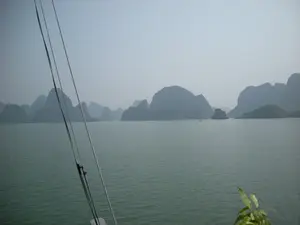 Bức ảnh chụp phong cảnh trên Vịnh Hạ Long. Khung cảnh bao gồm mặt biển rộng lớn màu xanh lục và rất nhiều hòn đảo đá vôi với các kích cỡ khác nhau nhô lên từ mặt nước. Bầu trời có sương mù, làm cho cảnh vật ở xa trở nên mờ ảo. Ở phía xa, có một chiếc thuyền nhỏ (ghe/tàu) đang di chuyển. Ở cận cảnh, có thể thấy các sợi dây cáp hoặc dây thừng của con tàu du lịch mà từ đó bức ảnh được chụp. Góc dưới bên phải của ảnh có một vài chiếc lá cây xanh. Không có thông tin dạng chữ viết nào trong ảnh.。（日本語訳）。ハロン湾の風景を撮影した写真です。風景には、広大な緑色の海面と、水面から突き出た様々な大きさの多くの石灰岩の島々が含まれています。空は霧がかかっており、遠くの景色がぼやけて見えます。遠方には、一隻の小さな船（小舟/船）が移動しています。近景では、写真が撮影された遊覧船のケーブルまたはロープが見えます。写真の右下隅には、いくつかの緑の木の葉があります。写真の中には、いかなる文字情報もありません。