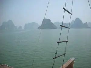 Bối cảnh là Vịnh Hạ Long, được chụp từ trên một chiếc tàu, có thể là tàu du lịch bằng gỗ. Ở phía trước, có thể thấy một phần của con tàu và một chiếc thang dây có các bậc bằng gỗ. Mặt nước biển rộng lớn, có màu xanh lục. Ở phía xa, nhiều hòn đảo đá vôi với thảm thực vật xanh tươi nhô lên từ mặt nước. Bầu trời có sương mù, tạo nên một khung cảnh mờ ảo. Hoạt động đang diễn ra là một chuyến du ngoạn trên vịnh.。（日本語訳）。背景はハロン湾で、船の上から撮影されたもので、おそらく木製の遊覧船でしょう。前景には、船の一部と、木製の踏み板がある縄梯子が見えます。広大な海面は緑色をしています。遠くには、青々とした植物に覆われた多くの石灰岩の島々が水面から突き出ています。空は霧がかかっており、幻想的な光景を作り出しています。行われている活動は、湾の遊覧です。
