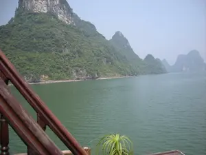 Khung cảnh được chụp từ trên một chiếc tàu du lịch đang di chuyển trên Vịnh Hạ Long. Mặt nước biển có màu xanh lục. Xung quanh là những hòn đảo đá vôi cao lớn, được bao phủ bởi cây cối xanh tươi. Dưới chân một hòn đảo gần nhất có một bãi biển nhỏ. Ở phía trước, góc dưới bên trái của bức ảnh là một phần lan can bằng gỗ của con tàu. Gần đó, ở góc dưới bên phải, có một chậu cây cảnh nhỏ. Ở phía xa, nhiều hòn đảo khác hiện ra mờ ảo trong sương mù, tạo nên một khung cảnh đặc trưng của Vịnh Hạ Long.。（日本語訳）。この風景は、ハロン湾上を移動している観光船の上から撮影されたものです。海水は緑色です。周りには、青々とした木々に覆われた、高くて大きな石灰岩の島々があります。最も近い島の麓には、小さなビーチがあります。その近く、右下隅には、小さな観葉植物の鉢があります。遠くには、他の多くの島々が霧の中にぼんやりと現れ、ハロン湾特有の風景を作り出しています。