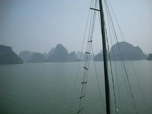 Bức ảnh chụp cảnh quan trên một vùng vịnh, rất có thể là Vịnh Hạ Long. Nổi bật là vô số các hòn đảo đá vôi với nhiều kích cỡ và hình dáng khác nhau, được bao phủ bởi cây xanh, nhô lên trên mặt biển màu xanh ngọc. Bầu trời có sương mù bao phủ, tạo nên một không gian huyền ảo và tĩnh lặng. Ở cận cảnh bên phải là một phần của cột buồm và hệ thống dây thừng của một chiếc thuyền, cho thấy bức ảnh được chụp từ một con tàu đang di chuyển trên vịnh, có thể là một chuyến du ngoạn ngắm cảnh. Không có bất kỳ chữ viết hay ký hiệu nào có thể đọc được trong ảnh.。（日本語訳）。ある湾の風景を撮影した写真で、おそらくハロン湾でしょう。目立っているのは、様々な大きさと形をした無数の石灰岩の島々で、緑樹に覆われ、エメラルドグリーンの海面から突き出ています。空は霧に覆われており、幻想的で静寂な空間を作り出しています。右側の前景には船の帆柱とロープ類の一部があり、この写真が湾上を移動中の船から撮影されたこと、おそらくは観光クルーズであろうことを示しています。写真の中には、読み取れる文字や記号は一切ありません。