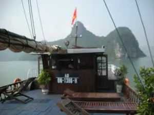 Bối cảnh là trên boong của một chiếc tàu du lịch bằng gỗ đang di chuyển trên Vịnh Hạ Long. Trên cabin bằng gỗ của tàu có ghi số hiệu đăng ký là "48" và "QN-1386-H". Trên nóc cabin có một cột cờ treo lá cờ đỏ sao vàng của Việt Nam. Boong tàu có sàn gỗ màu xanh, được trang bị một số ghế dài bằng gỗ để du khách tắm nắng và ngắm cảnh. Ngoài ra còn có các chậu cây cảnh và một chiếc phao cứu sinh màu cam. Ở phía sau, có thể thấy những hòn đảo núi đá vôi đặc trưng của Vịnh Hạ Long nổi lên trên mặt nước.。（日本語訳）。背景は、ハロン湾上を移動している木造遊覧船の甲板です。船の木造キャビンには、「48」と「QN-1386-H」という登録番号が記されています。キャビンの屋根の上には、ベトナムの金星紅旗を掲げた旗竿があります。甲板は青い木製の床で、観光客が日光浴をしたり景色を眺めたりするための木製長椅子がいくつか備え付けられています。その他にも、観葉植物の鉢やオレンジ色の救命浮環があります。後方には、ハロン湾特有の石灰岩の島々が水面に浮かび上がっているのが見えます。