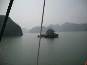 Khung cảnh được chụp từ trên một chiếc thuyền đang di chuyển trên biển, có khả năng là tại Vịnh Hạ Long. Phong cảnh đặc trưng với rất nhiều hòn đảo đá vôi lớn nhỏ nhô lên trên mặt nước. Bầu trời u ám và có sương mù, làm cho các hòn đảo ở phía xa trông mờ ảo. Ở phía trước, có thể thấy rõ một sợi dây thừng của con thuyền. Mặt biển khá tĩnh lặng, có màu xanh sẫm. Hoạt động đang diễn ra là một chuyến du ngoạn bằng thuyền.。（日本語訳）。海上を移動中の船から撮影された光景で、ハロン湾である可能性があります。風景は、水面から突き出た大小多数の石灰岩の島々が特徴です。空は曇って霧がかかっており、遠くの島々を霞ませています。手前には、船のロープが一本はっきりと見えます。海面はかなり穏やかで、濃い緑色をしています。行われている活動は、船での遊覧です。