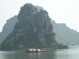 Bức ảnh chụp một cảnh quan đặc trưng của Vịnh Hạ Long, với những hòn đảo đá vôi hùng vĩ nhô lên từ mặt nước. Ở trung tâm và tiền cảnh là một hòn đảo đá lớn, được bao phủ bởi cây cối xanh tươi. Dưới chân đảo, một nhóm gồm khoảng ba, bốn chiếc thuyền nan nhỏ được buộc lại với nhau và đang nổi trên mặt nước. Những chiếc thuyền này có mái che hình vòm, có thể được làm bằng tre nứa và phủ bạt, và trên thuyền có cắm cờ đỏ. Đây có thể là những chiếc thuyền chở khách du lịch đi tham quan các hang động hoặc khu vực gần các đảo đá. Phía sau là các dãy núi đá vôi khác mờ dần trong làn sương, tạo nên một khung cảnh nhiều lớp. Mặt biển phẳng lặng, có màu xanh ngọc. Không có chữ viết nào có thể đọc được trong ảnh.。（日本語訳）。写真はハロン湾の典型的な景観を写しており、雄大な石灰岩の島々が水面から突き出ている。中央と前景には、青々とした木々に覆われた大きな岩の島がある。島の麓では、3、4隻ほどの小さな手漕ぎボートの集団が互いに結ばれ、水面に浮かんでいる。これらのボートにはアーチ型の屋根があり、それは竹でできて防水シートで覆われている可能性があり、ボートには赤い旗が立てられている。これらは、洞窟や岩島の近くのエリアを訪れる観光客を乗せるボートかもしれない。後方には、他の石灰岩の山々が霧の中にぼんやりと見え、多層的な風景を作り出している。海面は穏やかで、エメラルドグリーン色をしている。写真の中には読み取れる文字はない。