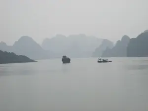 Bức ảnh chụp cảnh Vịnh Hạ Long trong một ngày có sương mù dày đặc. Khung cảnh chủ đạo là mặt biển rộng lớn, phẳng lặng và các dãy núi đá vôi trùng điệp ở phía xa, mờ ảo trong sương. Ở giữa vịnh, có một hòn đảo đá nhỏ đứng đơn độc. Về phía bên phải, một chiếc thuyền (ghe/tàu) có mái che đang di chuyển trên mặt nước, kéo theo một chiếc thuyền nhỏ hơn. Đây có thể là thuyền của ngư dân địa phương đang đi đánh cá hoặc là thuyền du lịch nhỏ. Phía bên trái có một phần của một hòn đảo lớn hơn. Không có chữ viết nào có thể nhận dạng được trong hình ảnh.。（日本語訳）。濃い霧が出た日のハロン湾の風景を撮影した写真です。湾の中央には、小さな岩の島が一つぽつんと立っています。右側には、屋根付きの船（小舟/ボート）が水面を移動しており、その後ろにさらに小さな船を引いています。これは、漁に出る地元の漁師の船か、あるいは小型の観光船かもしれません。左側には、より大きな島の一部が見えます。画像内には、認識できる文字はありません。