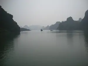 Bức ảnh chụp khung cảnh trên Vịnh Hạ Long. Bầu trời có sương mù nhẹ, tạo nên một không gian mờ ảo. Mặt nước vịnh khá phẳng lặng. Xung quanh là rất nhiều hòn đảo đá vôi lớn nhỏ với thảm thực vật xanh bao phủ, đặc trưng của khu vực này. Ở giữa vịnh, có thể thấy hai chiếc thuyền đang di chuyển: một chiếc tàu lớn hơn, có thể là tàu du lịch, và một chiếc thuyền nhỏ hơn ở phía xa. Hoạt động chính là sự di chuyển của các phương tiện đường thủy trên vịnh. Không có bất kỳ văn bản hay chữ viết nào có thể nhận dạng được trong ảnh.。（日本語訳）。ハロン湾の風景を撮影した写真です。空には薄い霧がかかっており、幻想的な空間を作り出しています。湾の水面はかなり穏やかです。周りには、この地域の特徴である緑の植生に覆われた大小さまざまな石灰岩の島々がたくさんあります。湾の中央では、2隻の船が移動しているのが見えます。1隻はより大きな船で、おそらく観光船でしょう。もう1隻は遠くにあるより小さな船です。主な活動は、湾上での水上交通機関の移動です。写真の中には、認識できるテキストや文字は一切ありません。