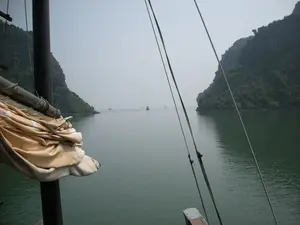 Bức ảnh được chụp từ trên một chiếc thuyền du lịch đang di chuyển trên Vịnh Hạ Long. Ở phía trước, có thể thấy một phần của con thuyền với cột buồm bằng gỗ, một cánh buồm màu vàng đã được cuộn lại và các dây thừng. Con thuyền đang đi qua một eo biển với mặt nước phẳng lặng màu xanh lục. Hai bên là những hòn đảo đá vôi cao lớn, được bao phủ bởi cây cối xanh um. Ở phía xa, có thể nhìn thấy một vài chiếc thuyền khác đang lướt trên mặt vịnh. Bầu trời có màu xám nhạt và hơi có sương mù, tạo nên một khung cảnh yên bình và đặc trưng của vùng vịnh. Không có văn bản hay chữ viết nào trong ảnh.。（日本語訳）。この写真は、ハロン湾を移動中の観光船の上から撮影されたものです。前方には、木製のマスト、巻き上げられた黄色い帆、そしてロープなど、船の一部が見えます。船は、緑色で穏やかな水面の海峡を通過しています。両側には、青々とした木々に覆われた、高くて大きな石灰岩の島々があります。遠くには、湾の水面を滑るように進む他の船が数隻見えます。空は薄い灰色で、少し霧がかかっており、この湾の地域に特有の穏やかな風景を作り出しています。写真にはテキストや文字はありません。