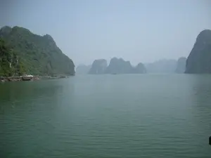 Bức ảnh chụp phong cảnh Vịnh Hạ Long. Mặt biển rộng lớn, phẳng lặng có màu xanh ngọc. Xung quanh là rất nhiều hòn đảo đá vôi với các kích cỡ và hình dạng khác nhau, được bao phủ bởi thảm thực vật xanh tươi. Bầu trời có sương mù bao phủ, làm cho cảnh vật ở xa trở nên mờ ảo. Ở phía xa, một chiếc tàu nhỏ đang di chuyển trên mặt nước. Dọc theo chân của hòn đảo lớn bên trái, có thể thấy một dãy các công trình nổi trên mặt nước, có khả năng là một làng chài hoặc các bè nuôi trồng thủy sản.。（日本語訳）。ハロン湾の風景を撮った写真です。広大で穏やかな海面は、エメラルドグリーン色をしています。周りには、様々な大きさや形の石灰岩の島々がたくさんあり、青々とした植物に覆われています。空は霧に覆われており、遠くの景色がぼんやりと見えます。遠くには、一隻の小さな船が水面を移動しています。左側の大きな島の麓に沿って、水上に浮かぶ一連の建造物が見えます。それは漁村か水産養殖用のいかだである可能性があります。