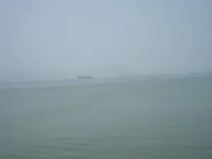 Khung cảnh trên biển, có thể là Vịnh Hạ Long, vào một ngày có sương mù dày đặc. Mặt nước biển có màu xanh xám và khá tĩnh lặng. Sương mù bao phủ làm cho tầm nhìn bị hạn chế. Ở phía xa, có thể nhìn thấy bóng của một con tàu lớn, có thể là tàu chở hàng, đang ở trên mặt biển. Xa hơn nữa, ẩn hiện trong làn sương là bóng dáng của những hòn đảo hoặc núi đá vôi.。（日本語訳）。濃い霧の日の、おそらくハロン湾と思われる海上の景色。海面は灰色がかった青色で、かなり穏やかです。霧が覆っているため、視界が制限されています。遠方には、おそらく貨物船と思われる大きな船の影が海上にあります。さらにその奥、霧の中に島々や石灰岩の山の影が見え隠れしています。