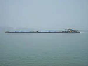 Trong ảnh là một chiếc sà lan (ghe/tàu) rất dài đang di chuyển trên mặt biển. Bối cảnh là một vùng biển rộng lớn, có thể là Vịnh Hạ Long, vào một ngày có sương mù hoặc thời tiết mờ ảo, làm cho tầm nhìn bị hạn chế. Chiếc sà lan đang vận chuyển hàng hóa, được che phủ cẩn thận bằng các tấm bạt lớn màu xanh dương. Phần đuôi của sà lan là một cabin điều khiển và động cơ, có chức năng đẩy toàn bộ sà lan về phía trước. Hoạt động chính đang diễn ra là vận tải hàng hóa đường thủy. Ở phía xa, qua lớp sương mù, có thể thấy lờ mờ hình ảnh của đất liền với một vài công trình xây dựng. Không có chữ viết nào có thể đọc được trên thân tàu.。（日本語訳）。背景は広大な海域で、おそらくハロン湾でしょう。霧または霞んだ天候の日で、視界が制限されています。はしけは貨物を輸送しており、大きな青いシートで慎重に覆われています。はしけの後部は操縦室とエンジンで、はしけ全体を前方に押し出す機能を持っています。行われている主な活動は水上貨物輸送です。遠くには、霧を通して、いくつかの建設中の建物がある陸地の姿がぼんやりと見えます。船体には読み取れる文字はありません。