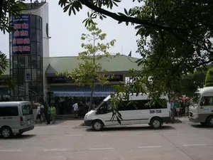 Đây là hình ảnh chụp tại Cảng tàu khách du lịch Bãi Cháy, một nhà ga cảng biển phục vụ các chuyến tham quan Vịnh Hạ Long.。**Chữ viết được ghi lại:**。*   Trên biển hiệu lớn màu xanh dương treo trên tòa tháp của tòa nhà có ghi: "CẢNG TÀU KHÁCH DU LỊCH BÃI CHÁY" và "TRUNG TÂM ĐIỀU HÀNH TÀU THAM QUAN VỊNH HẠ LONG".。*   Phía trên lối vào tòa nhà có biển hiệu "PHÒNG BÁN VÉ" và chữ "TICKET".。*   Một băng rôn treo dưới mái hiên có dòng chữ: "DU LỊCH VỊNH HẠ LONG VÌ MỘT MÔI TRƯỜNG XANH".。**Mô tả cảnh quan và hoạt động:**。*   **Loại địa điểm:** Đây là một nhà ga cảng tàu du lịch, cụ thể là Cảng tàu khách Bãi Cháy, nơi có phòng bán vé và trung tâm điều hành các tour tham quan Vịnh Hạ Long.。*   **Hoạt động cụ thể:** Nhiều du khách đang tập trung trước cửa tòa nhà. Một số người đang đi lại, trong khi những người khác đang đứng thành nhóm, có vẻ như đang chờ đợi để mua vé hoặc chờ hướng dẫn viên. Ở phía bên phải, một nhóm du khách, trong đó có người đội nón lá, đang đứng trên vỉa hè gần một chiếc xe buýt nhỏ. Hoạt động chung cho thấy các du khách đang chuẩn bị cho chuyến đi tham quan Vịnh Hạ Long bằng tàu.。*   **Vật thể trong ảnh:** Tòa nhà chính là một nhà ga hiện đại với một tòa tháp bằng kính. Phía trước là một con đường hoặc bãi đỗ xe có một vài chiếc xe ô tô, bao gồm xe van và xe buýt nhỏ, dường như là xe chở khách du lịch. Có nhiều cây xanh lớn che bóng mát xung quanh khu vực.。（日本語訳）。これはバイチャイ旅客船港で撮影された写真です。ハロン湾観光ツアーに利用される海港ターミナルです。**記録された文字：**。*   建物の塔に掲げられた大きな青い看板には、「CẢNG TÀU KHÁCH DU LỊCH BÃI CHÁY」（バイチャイ旅客船港）と「TRUNG TÂM ĐIỀU HÀNH TÀU THAM QUAN VỊNH HẠ LONG」（ハロン湾観光船運航センター）と書かれています。*   建物の入口の上には、「PHÒNG BÁN VÉ」（チケット売り場）と「TICKET」という文字の看板があります。*   軒下に掲げられた横断幕には、「DU LỊCH VỊNH HẠ LONG VÌ MỘT MÔI TRƯỜNG XANH」（環境に優しいハロン湾観光のために）という文言が書かれています。**風景と活動の説明：**。*   **場所の種類：**これは旅客船港のターミナルで、具体的にはバイチャイ旅客船港です。ここにはチケット売り場とハロン湾観光ツアーの運航センターがあります。*   **具体的な活動：**多くの観光客が建物の前に集まっています。歩き回っている人もいれば、グループで立っている人もおり、チケットの購入を待っているか、ガイドを待っているようです。右側では、ノンラー（ベトナムの伝統的な笠）をかぶった人を含む観光客のグループが、小さなバスの近くの歩道に立っています。全体的な活動から、観光客が船でのハロン湾観光の準備をしていることがわかります。*   **写真の中の物体：**主要な建物はガラス張りの塔を持つ近代的なターミナルです。その前には道路または駐車場があり、バンや小型バスを含む数台の車があります。これらは観光客を運ぶ車のようです。周辺には日陰を作る大きな緑の木々がたくさんあります。