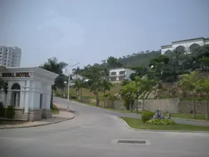 Bức ảnh chụp một con đường nhựa trong một khu vực có vẻ là khu nghỉ dưỡng hoặc đô thị cao cấp, có thể ở Hạ Long. Bên trái là một công trình kiến trúc màu trắng giống như cổng chào, trên đó có dòng chữ 'ROYAL HOTEL' màu vàng. Phía sau công trình này là một tòa nhà cao tầng hiện đại. Con đường vắng vẻ, không có người hay phương tiện giao thông. Bên phải con đường là một sườn đồi được bao phủ bởi cây xanh và có một vài biệt thự màu trắng được xây dựng trên đó. Dọc theo sườn đồi là một bức tường đá. Nhiều cây cọ được trồng dọc hai bên đường. Ở góc đường có một tiểu cảnh trang trí bằng đá và cây bụi. Bầu trời trong ảnh có màu xám nhạt và hơi mờ.。（日本語訳）。写真は、おそらくハロンにある、リゾート地または高級都市部と思われる地域のアスファルトの道を写しています。左側にはウェルカムゲートのような白い建築物があり、その上には黄色い文字で「ROYAL HOTEL」と書かれています。この建築物の後ろには、近代的な高層ビルがあります。道は閑散としており、人や乗り物はありません。道の右側は緑樹に覆われた丘の斜面で、その上にはいくつかの白いヴィラが建てられています。丘の斜面に沿って石垣があります。道の両脇には多くのヤシの木が植えられています。道の角には、石と低木で装飾された小さな造園があります。写真の空は薄い灰色で、少し霞んでいます。